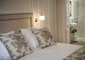 A neatly made bed with floral-patterned pillows, a wall-mounted light fixture, and a glimpse of a bathroom with a sink and shower.