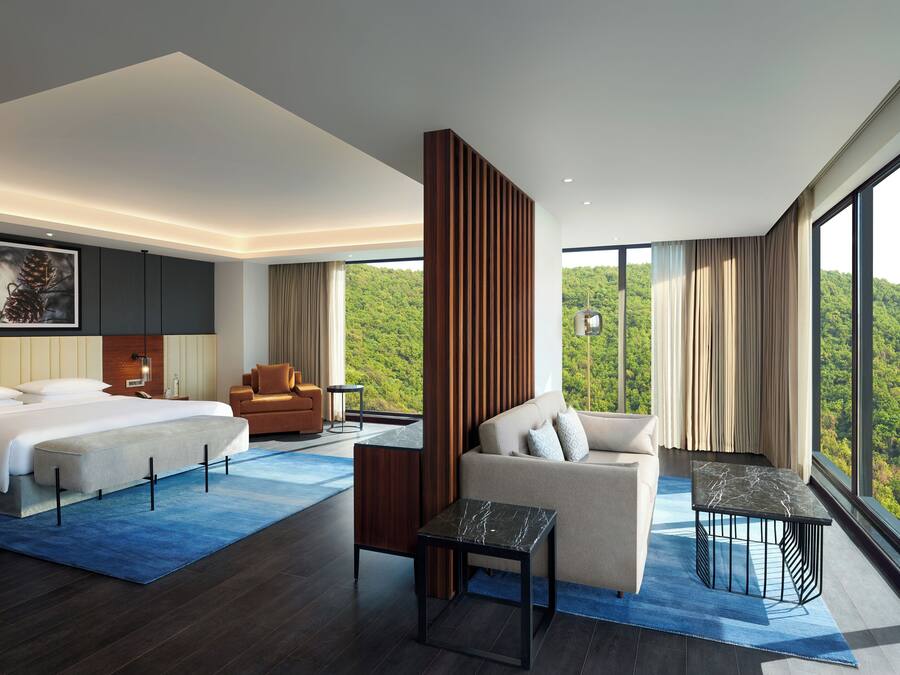 A modern hotel room with a large bed, a sofa, a small table, and a view of a forest.