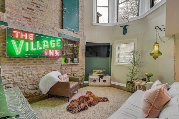 A cozy living room with a brick wall, a neon sign reading "The Village Inn", a fireplace, and a large window.