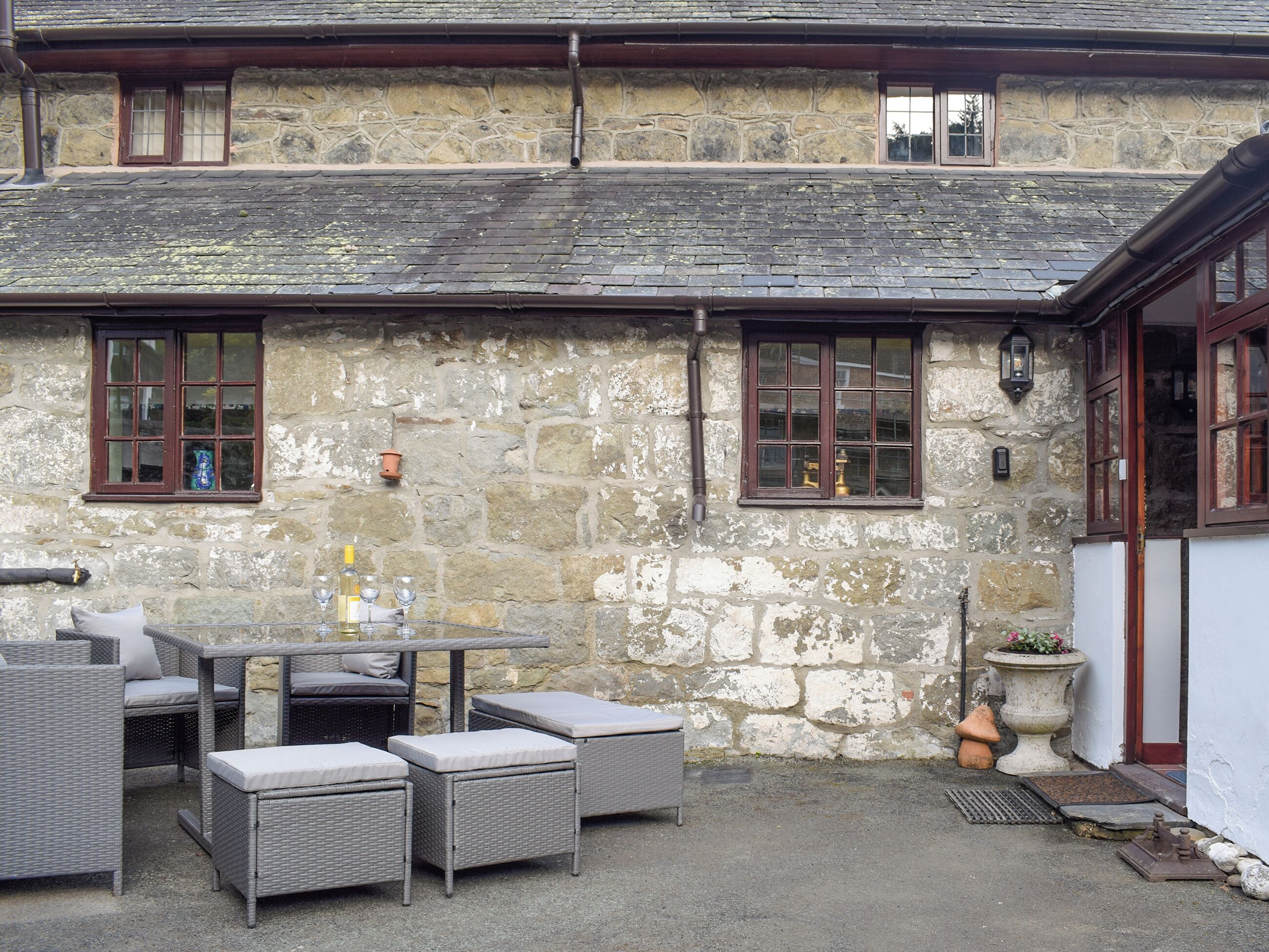 A stone building with a tiled roof, outdoor seating with wicker furniture, and a small garden area with a flower pot.