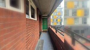 Un corridoio stretto con un muro di mattoni su un lato e una ringhiera metallica sull'altro. In lontananza, si intravede una porta verde.