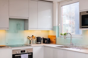 A modern kitchen with white cabinets, a stainless steel sink, a coffee maker, and a window with a view of buildings. - Spacious 3BD Duplex Steps from Old Street Station (London)