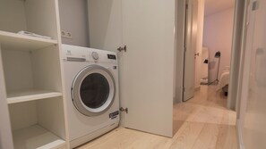 A compact laundry room with a front-loading washing machine, a shelf above it, and a door leading to a bathroom.