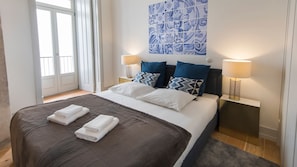 Un lit bien fait, recouvert d’une couverture marron, avec des oreillers blancs et deux serviettes pliées. Deux lampes de chevet sont placées de chaque côté du lit.