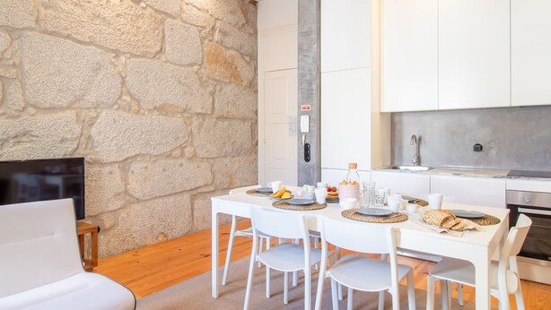 A modern kitchen with white cabinets, a stone wall, and a dining area with white chairs and a table set for breakfast. - Dirk Apartment - Almada 3º T by LovelyStay (Porto)