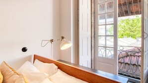 A bedroom with a wooden headboard, a bedside lamp, and a window with a view of a balcony and greenery. - Dirk Apartment - Almada 3º T by LovelyStay (Porto)