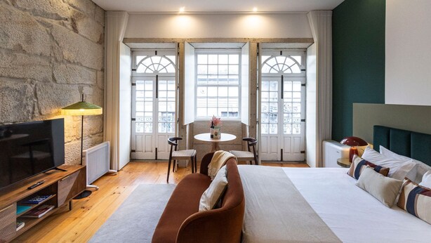 A hotel room with a stone wall, a bed with a headboard, a TV, a table with chairs, and large windows with a view. - Allderman Apartment - Almada 3º F by LovelyStay (Porto)