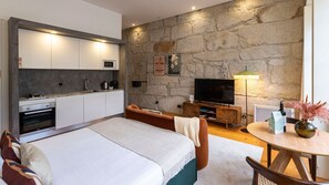 A modern bedroom with a stone wall, a flat-screen TV, a dining area, and a kitchenette. - Allderman Apartment - Almada 3º F by LovelyStay (Porto)