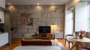 A modern living room with a stone wall, a flat-screen TV, a sofa, a dining table, and a kitchen area with a microwave. - Allderman Apartment - Almada 3º F by LovelyStay (Porto)