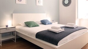 Un letto rifatto con cuscini, una lampada da comodino e un asciugamano piegato su un comodino bianco.