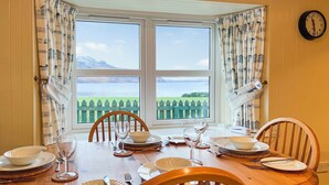 A dining table set for four with chairs, a clock on the wall, and a view of a body of water through a window. - 3 bedroom accommodation in Arnisdale, Kyle of Lochalsh (Arnisdale, Kyle of Lochalsh)