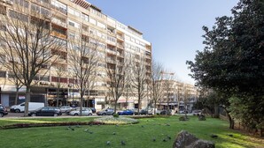 Um edifício de vários andares com uma fileira de lojas no andar térreo, uma área gramada com pombos e carros estacionados.