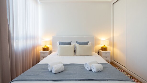 Quarto de hotel com cama, duas mesas de cabeceira, duas luminárias, um cobertor cinza e duas toalhas enroladas sobre a cama.