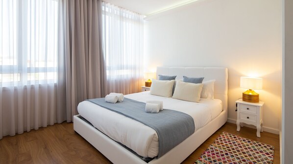 Une chambre d’hôtel moderne avec un grand lit, du linge de lit blanc, deux lampes de chevet, une table de chevet et un tapis coloré.