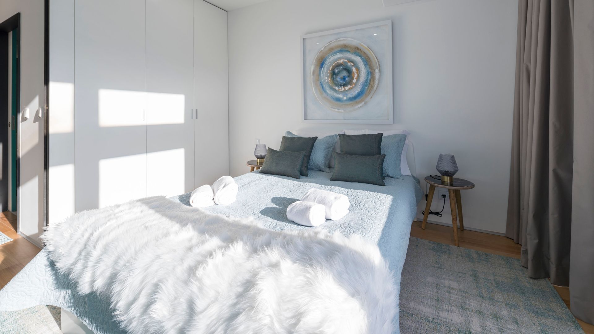 Un dormitorio moderno con una cama grande, varios almohadones, un plaid blanco suave, una mesita de noche con lámpara y un cuadro en la pared.