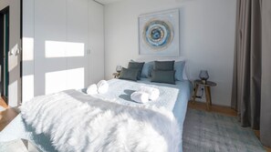 A modern bedroom with a large bed, multiple pillows, a fluffy white throw, a side table with a lamp, and a painting on the wall.