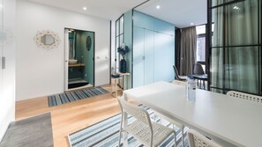 A modern hotel room with a large window, a white dining table, and a separate bathroom area.