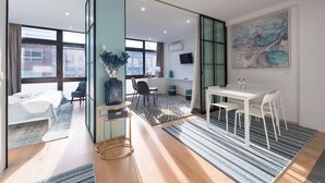 A modern hotel room with a large bed, a sitting area with a small table and chairs, a dining area with a white table and chairs, and a large window with a view of the city. - Wohaza Flat w/ Parking by LovelyStay (Porto)