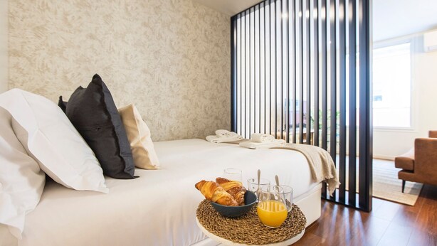 Ein ordentlich bezogenes Bett mit Kissen, ein Nachttisch mit Orangensaft und Croissants, und ein Blick ins Freie durch vertikale Jalousien.
