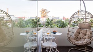 Ein Balkon mit einem runden Tisch, zwei weißen Stühlen, einem Hängesessel und Blick auf Gebäude und Bäume.