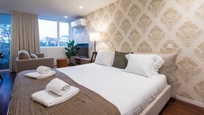 Ein Hotelzimmer mit einem großen Bett, einem Schreibtisch mit Computer, einem Sessel und Blick auf die Stadt durch das Fenster.