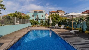 Ein Schwimmbecken mit Holzterrasse, umgeben von Grünpflanzen und einem Zaun.