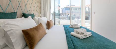 A hotel room with a bed, pillows, a teal throw blanket, and a view of the city through large windows.