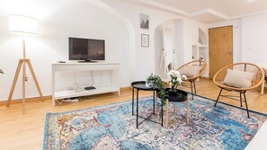 Una sala de estar moderna con un televisor de pantalla plana, un mueble blanco para entretenimiento, sillas de mimbre, una alfombra estampada y suelo de madera.