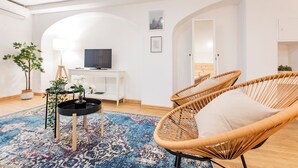 Un salon moderne avec un grand tapis, des fauteuils en osier, une console blanche et un téléviseur.