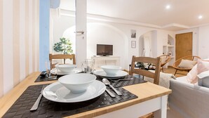 Un coin repas avec une table en bois dressée pour quatre personnes, des assiettes blanches, des bols et des couverts. Au fond, on aperçoit un téléviseur posé sur un meuble, une chaise avec un coussin et une plante sur le rebord de la fenêtre.