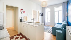 A modern kitchen with white cabinetry, a marble countertop, and a wooden bowl. There is a dining area with a blue sofa and a TV mounted on the wall.