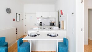 A compact kitchen with white cabinets, a coffee machine, and a microwave. There is a dining table with two chairs and a small island with a bowl and wine glasses. - Porto Flores Plaza by LovelyStay (Porto)