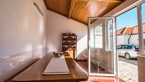 Una habitación iluminada por el sol con un banco de madera, una estantería con vajilla y vista a un coche a través de un umbral abierto.