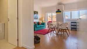 Ein modernes Wohnzimmer mit einem petrolfarbenen Sofa, einem Holzespeisetisch und Stühlen, einem roten Teppich und einem großen Fenster mit Blick.