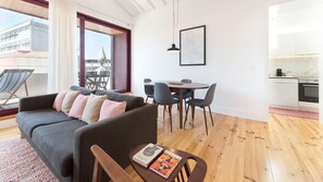 A modern living room with a grey sofa, a dining table with chairs, a kitchen area, and a large window with a view of buildings.