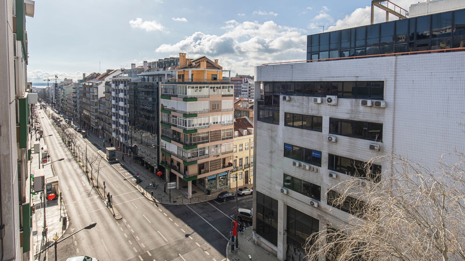 A multi-story building with a modern glass facade, adjacent to a street with parked cars and a pedestrian crossing.