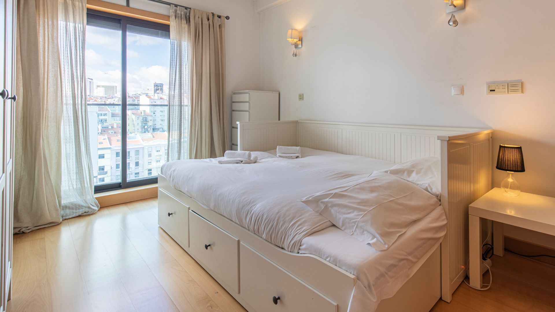 A hotel room with a large window, a bed with white linens, a bedside table with a lamp, and a dresser with drawers.