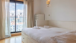 Una habitación de hotel con un ventanal grande, una cama con sábanas blancas, una mesita de noche y una lámpara de pared.