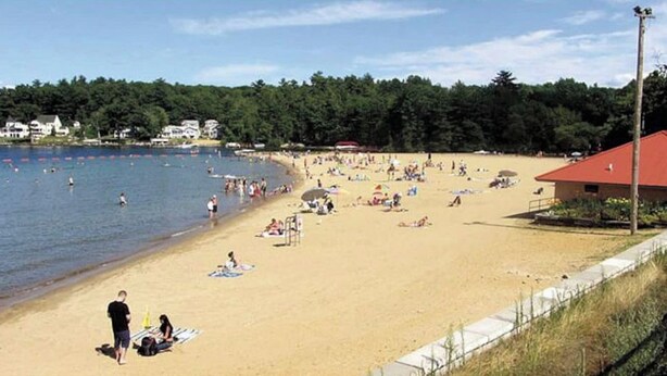Une plage où des gens se prélassent au soleil et se baignent, un bâtiment avec un toit rouge et, au fond, une zone boisée.