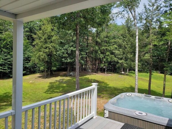 A hot tub on a wooden deck with a white railing, surrounded by a lush green lawn and tall trees.