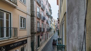 Eine schmale Straße, gesäumt von mehrstöckigen Gebäuden, einige mit Balkonen und Schildern für ein Bistro und eine Bar.