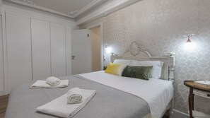 A neatly made bed with white linens and two pillows, a white wardrobe, and a small bedside table with a lamp.
