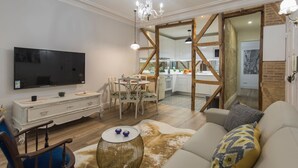A modern living room with a flat-screen TV, a dining area, and a kitchen visible through an open doorway.
