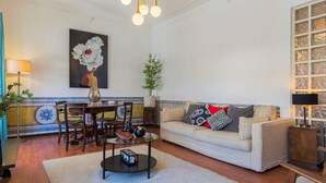 A living room with a beige sofa, a glass coffee table, a dining table with chairs, a painting on the wall, and a potted plant. - Charming Chiado 2BDR W/Balcony by LovelyStay (Encarnação)