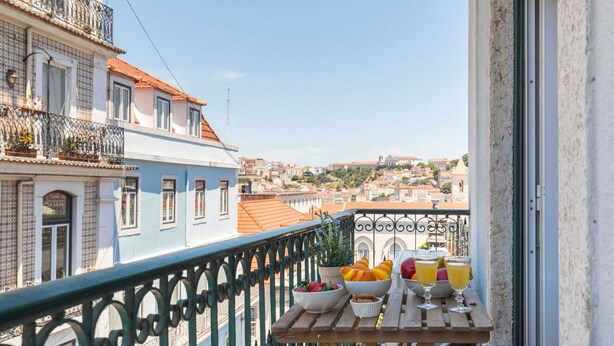 A balcony with a view of a cityscape, a table with fruit and drinks, and a railing. - Charming Chiado 2BDR W/Balcony by LovelyStay (Encarnação)