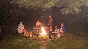 A group of people sitting around a campfire in an outdoor setting with trees in the background.