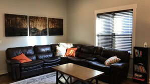 A living room with a black leather sectional sofa, a wooden coffee table, and a patterned rug. There are three wall-mounted paintings, a window with blinds, and a bookshelf with books.