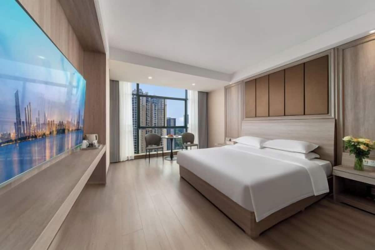 Une chambre d’hôtel moderne dotée d’un grand lit, d’un coin salon avec deux fauteuils, d’une grande fenêtre offrant une vue sur la ville et d’un téléviseur fixé au mur.