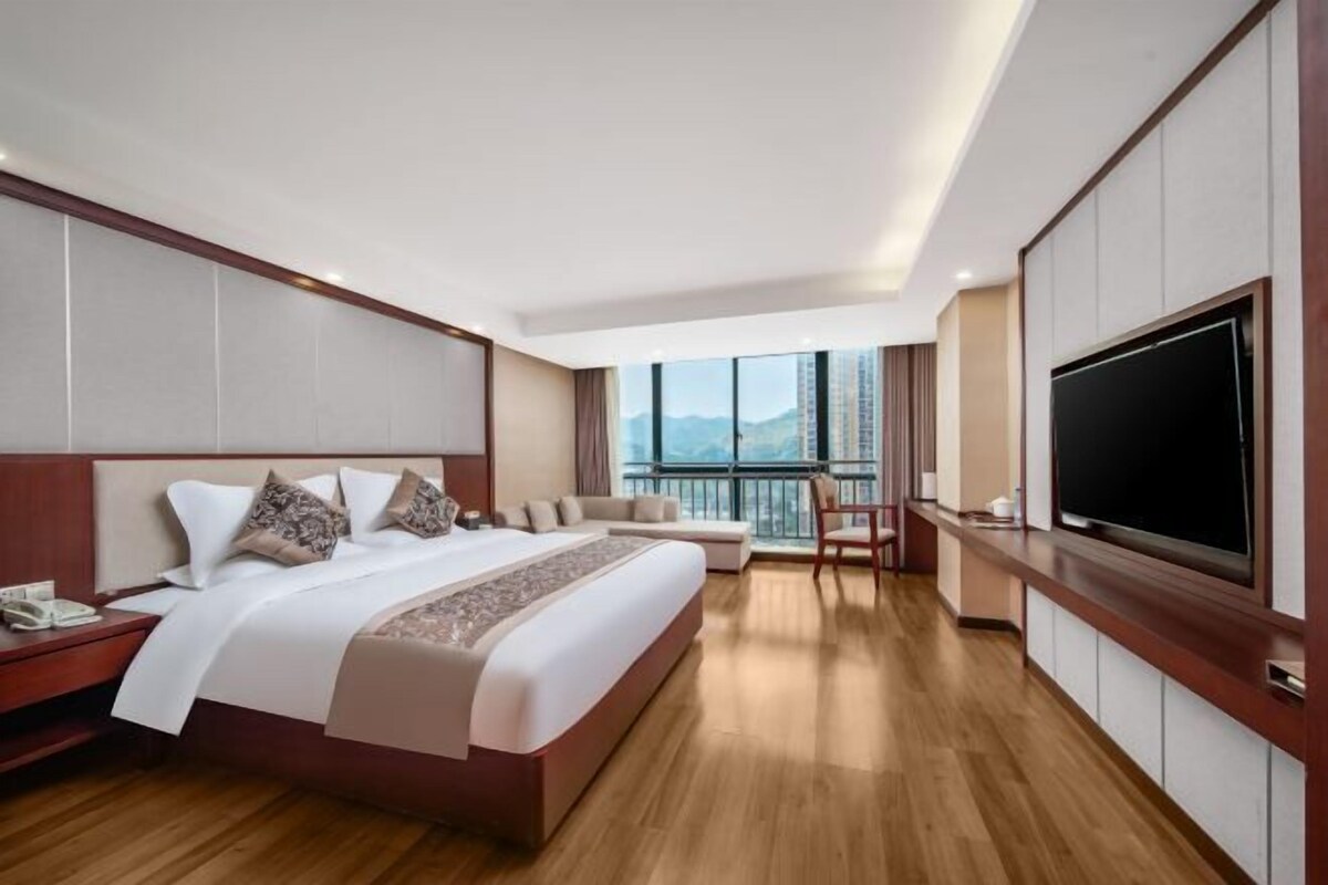 A modern hotel room with a large bed, a TV, a balcony with a city view, and a wooden floor.