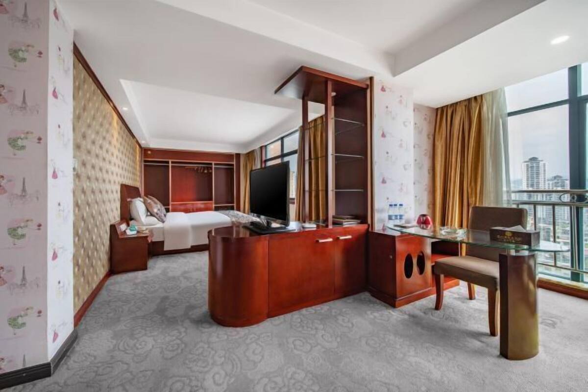 Ein Hotelzimmer mit Bett, Fernseher, Schreibtisch mit Stuhl, Kleiderschrank und großem Fenster mit Blick auf die Stadt.